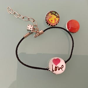 Puede incluir: Una pulsera de cordón negro con una cadena plateada y un dije de dado. La pulsera presenta un dije redondo con un corazón rosa y la palabra "Love". También hay dos botones decorativos.