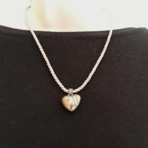 Peut inclure: Collier argenté avec un pendentif en forme de cœur. Le pendentif présente une surface texturée et est suspendu à une chaîne torsadée. Le collier est présenté sur un fond noir.