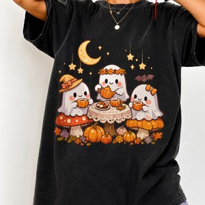 Peut inclure: Un t-shirt noir à manches courtes avec un motif représentant trois fantômes blancs assis à une table, buvant dans des tasses orange. La scène comprend un croissant de lune, des étoiles, une chauve-souris, des citrouilles et des champignons.