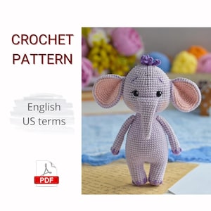 Puede incluir: Un elefante de ganchillo lavanda con grandes orejas de color rosa claro y una pequeña flor morada en la cabeza. La imagen incluye el texto "CROCHET PATTERN" y "English US terms". También se ve un icono PDF.