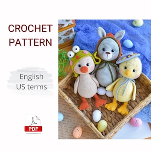 Op de afbeelding: Haakpatroon met drie handgemaakte amigurumi-speeltjes: een eend met een kikkerhoed, een konijn en een kuiken met een zonnehoed. De speeltjes liggen in een gevlochten mand met paaseieren. De tekst "CROCHET PATTERN" en "English US terms" zijn ook zichtbaar.