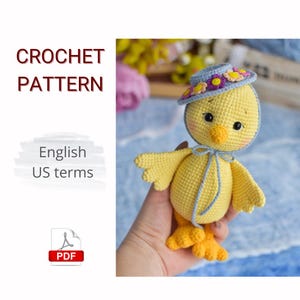 May include: A crocheted yellow chick wearing a blue hat with flower accents. The chick has a yellow beak, black eyes, and blue bow. The image includes the text "CROCHET PATTERN" and "English US terms". A PDF icon is also present.