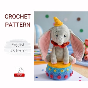 Może przedstawiać: Szydełkowa zabawka w kształcie słonia z dużymi uszami wyłożonymi różem, żółtym kapeluszem i czerwonym kołnierzem, siedząca na żółto-niebieskiej platformie w stylu cyrkowym. Obraz zawiera tekst "CROCHET PATTERN" i "English US terms".