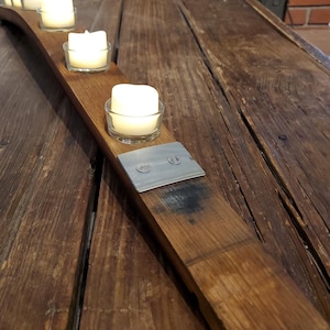 May include: A wooden candle holder made from a repurposed barrel stave. It features five clear glass votive holders with lit candles. The wood is a warm brown color, with metal accents.