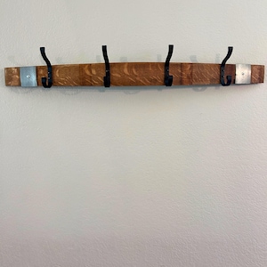 May include: A wooden coat rack with four black metal hooks. The rack is made from a section of a barrel, with visible wood grain and metal accents. The hooks are evenly spaced along the length of the rack, which is mounted on a white wall.