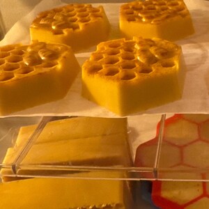 Puede incluir: Varios jabones amarillos con forma de panal de abeja están colocados en un estante. Los jabones tienen una superficie texturizada con un patrón de celdas hexagonales. Debajo, hay más barras de jabón rectangulares y un molde rojo y amarillo.