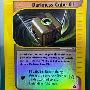 Puede incluir: Una carta de juego de Pokémon con un cubo metálico con antenas, etiquetada como "Darkness Cube 01". La tarjeta tiene un borde amarillo y el texto "Trainer" y "Technical Machine".