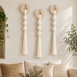 May include: Three wall hangings featuring light wood circular tops with open centres, connected to long, off-white macrame cords with spherical knots and finished with long, straight tassels hanging vertically against a neutral wall.