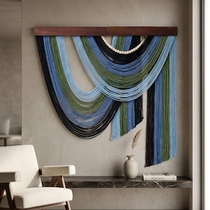 May include: A wall hanging featuring multiple strands of yarn in shades of black, olive green, and light blue, suspended from a horizontal dark wood bar. The yarn is arranged in draped, curved layers with a row of light-colored wooden beads.