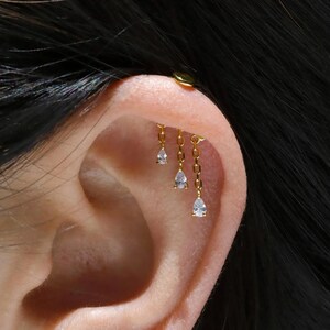 May include: A gold-toned cartilage earring featuring three dangling chains of varying lengths, each tipped with a small, clear, pear-shaped cubic zirconia stone. The earring is secured to the upper ear cartilage with a flat, circular gold-toned stud.