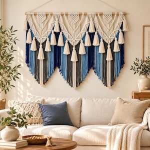 May include: A large macrame wall hanging suspended from a wooden rod, featuring intricate knotted patterns in cream cord. The design includes multiple hanging tassels and long, vertical fringe strands in a gradient of dark blue, light blue, and grey.