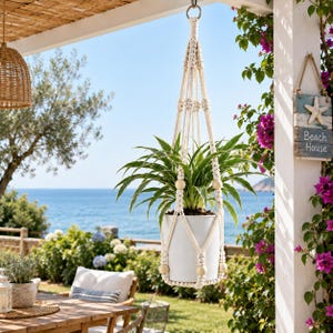 Puede incluir: Un colgador de plantas de macrame en color crema con intrincados patrones de cuerda anudada y cuentas de madera lisas. El soporte sostiene una maceta cilindrica blanca con una planta cinta verde, creando un elemento decorativo para el hogar.