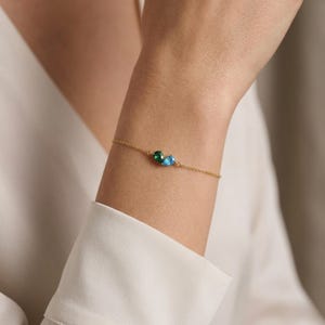 May include: A delicate gold bracelet featuring two gemstones: a green emerald and a blue topaz. The bracelet is worn on a wrist, showcasing the fine chain and the vibrant colors of the stones. Jewelry.
