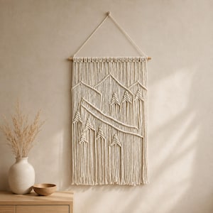 May include: A cream-coloured macrame wall hanging featuring a mountain range and forest scene created with intricate knotted cord patterns. The piece is suspended from a horizontal wooden dowel with a single cord loop for mounting on a wall.