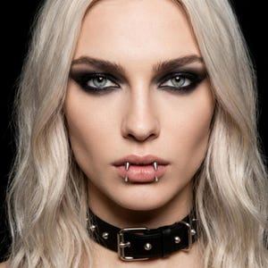 Könnte beinhalten: Ein Lippenpiercing aus silberfarbenem Metall mit zwei spitzen Dornen, die aus der Unterlippe ragen, kombiniert mit einem schwarzen Lederhalsband, das mit kleinen silberfarbenen Metallnieten und einer mittigen Schnalle versehen ist.