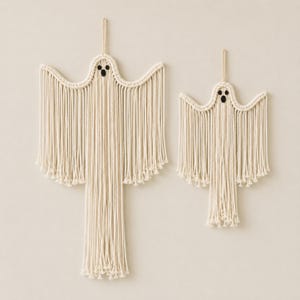May include: Two off-white macrame wall hangings shaped like ghosts. Each ghost features a knotted head with two small black circular eyes and one black oval mouth, with long vertical fringe strands extending downward to form the body.