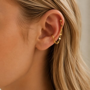 Op de afbeelding: Een goudkleurige oorbel bestaande uit een ear cuff verbonden met een kleine knop door een fijne ketting met platte, ronde schakels. De ear cuff zit op het buitenste kraakbeen, terwijl de knop hoger is geplaatst voor een kettingeffect over het oor.