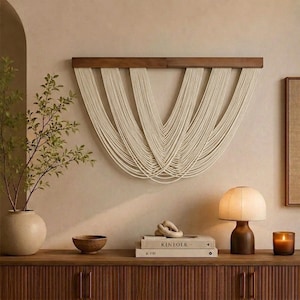 May include: A wall hanging featuring off-white cotton cords draped in multiple U-shaped layers from a horizontal dark brown wooden bar. The cords are arranged in a symmetrical pattern, creating a series of cascading loops that hang against the wall.