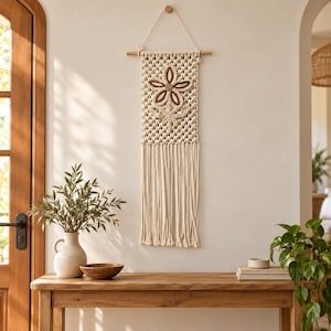 May include: A cream-colored macrame wall hanging featuring a central flower design with brown and tan accents. The piece is suspended from a wooden dowel with a hanging cord and finished with long, straight fringe at the bottom edge.