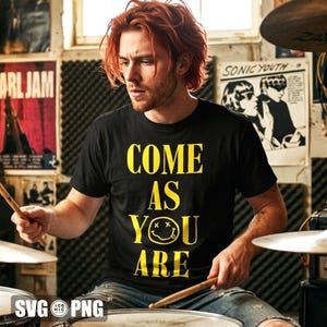 Op de afbeelding: Een zwart t-shirt met de gele tekst "COME AS YOU ARE" en een smiley-gezicht. De persoon houdt drumstokken vast en zit achter een drumstel. Posters zijn zichtbaar op de achtergrond.