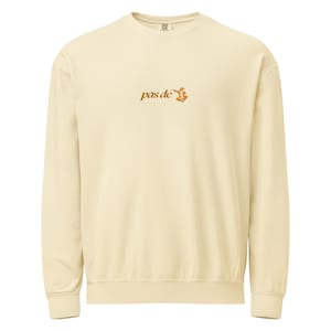 May include: Cream-colored sweatshirt with long sleeves. The text "pas de" is embroidered in a brown script, accompanied by a small orange illustration. The sweatshirt has a crew neck and ribbed cuffs and hem.