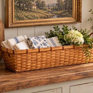 May include: A long, rectangular woven basket made of light brown wood strips with two small handles on the ends. The basket contains several folded fabric napkins with blue and white striped or floral patterns and a cluster of green leaves and white flowers.