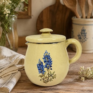 May include: A pale yellow ceramic jar with a matching lid and a single handle. The front of the jar features a detailed botanical illustration of blue lupine flowers with green stems and leaves, set against the speckled cream-colored glaze.