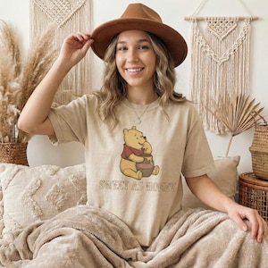 Puede incluir: Camiseta beige con un dibujo de Winnie the Pooh comiendo miel de un tarro. El texto "SWEET AS HONEY" está debajo del dibujo. La persona de la imagen lleva un sombrero marrón.