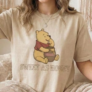 Puede incluir: Camiseta beige con una ilustración de dibujos animados de un oso con una camisa roja, sentado junto a un tarro de miel. El texto "SWEET AS HONEY" está impreso debajo en marrón. La camiseta tiene mangas cortas.