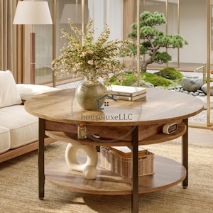 May include: A round, two-tiered coffee table with a dark brown wooden top and a black metal frame. The table has a lower shelf and a drawer. A vase of flowers and books sit on top. The text "houseluxeLLC" is visible.