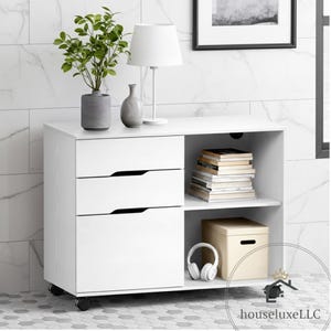 May include: A white storage cabinet with three drawers and an open shelf. The cabinet has a lamp, a vase, and a plant on top. The open shelf holds books, a box, and headphones. The cabinet is on wheels.