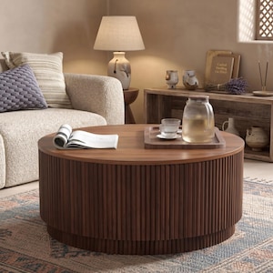 May include: A round, dark brown wooden coffee table with a vertical slatted base. The tabletop is flat and circular, featuring an open book, a square wooden tray, a clear glass pitcher filled with liquid, and two small glass cups on the surface.