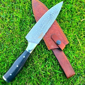 Puede incluir: Un cuchillo de chef de acero de Damasco con mango negro y virola plateada, sobre una funda de cuero marrón. La hoja tiene un patrón distintivo. El cuchillo y la funda están sobre hierba verde.