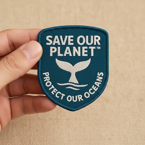 Könnte beinhalten: Ein tealfarbener Aufnäher mit den Worten "SAVE OUR PLANET" und "PROTECT OUR OCEANS" in Weiß. Ein weißes Walflossendesign ist auf dem Aufnäher zentriert. Der Aufnäher wird von einer Hand gehalten.