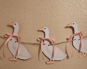 May include: A garland of five wooden goose cutouts. Each goose is painted white with a yellow beak and feet. A pink ribbon is tied around each goose's neck. The geese are strung on a natural twine.