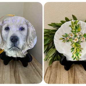 Puede incluir: Dos adornos decorativos de concha. Uno tiene forma de cabeza de perro, con la cara de un golden retriever. El otro es una concha con un diseño de cruz pintada, con flores blancas y hojas verdes. Ambos están sobre soportes negros.