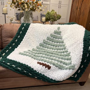 May include: A chunky knit blanket featuring a white background with a large, centered Christmas tree design in shades of sage green and white. The blanket has a thick, dark forest green border with white stitching accents along the edges.