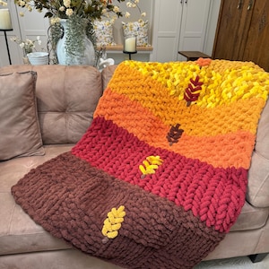 May include: A chunky knit throw blanket featuring a horizontal color-blocked design in shades of yellow, orange, red, and brown. The blanket is decorated with small, embroidered leaf-shaped appliques in contrasting colors placed along the stripes.
