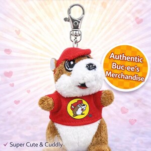 May include: A plush Buc-ee's keychain depicting a beaver character in a red shirt and hat. The keychain has a silver clasp and the text "Authentic Buc-ee's Merchandise". The image also includes the text "Super Cute & Cuddly".