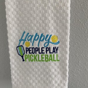 May include: A white waffle-weave hand towel featuring an embroidered design with the text Happy PEOPLE PLAY PICKLEBALL. The design includes a blue script font, dark blue block letters, and a green pickleball paddle with two yellow balls.