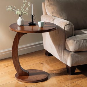 May include: A dark brown, C-shaped side table with a round top and base. A white vase with flowers, a black candle holder with a white candle, and a light blue mug sit on the table. The table is next to a beige sofa.