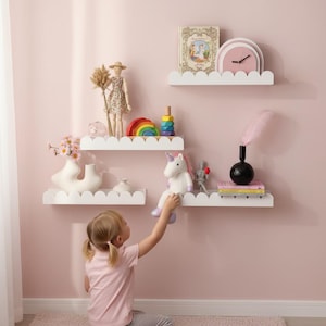 Op de afbeelding: Drie witte planken met geschulpte randen aan een roze muur tonen kinderspeelgoed en decoraties. Items zijn onder meer een pop, regenboogstapelaar, eenhoornknuffel, klok, boeken en vazen. Een jong kind reikt naar een speeltje.
