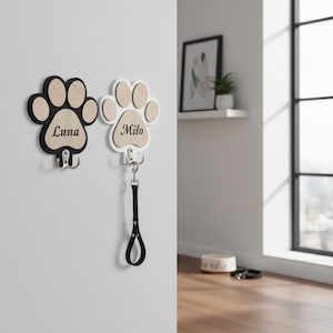 May include: Two wall-mounted paw print hooks made of wood and metal. One hook is black with the name Luna engraved, and the other is white with the name Milo engraved. Each paw print features a central hook for hanging items like a pet leash.