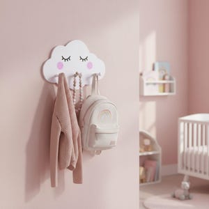 Op de afbeelding: Een witte, wolkvormige wandhaak met roze accenten en wimpers, met een roze trui, een witte rugzak met een regenboogontwerp en een kralenketting. De items hangen tegen een roze muur, wat wijst op een kinderkamerthema.