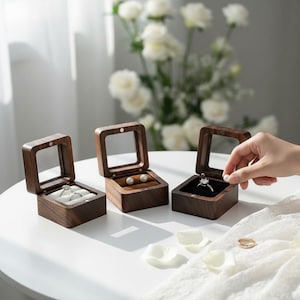 May include: Three wooden jewelry boxes with clear lids are open, displaying a ring, earrings, and a ring. The boxes are brown with a square shape. One box has a hand placing a pearl inside. The background includes white flowers.