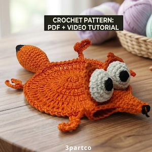 Op de afbeelding: Een oranje gehaakte vos met grote, witte en zwarte ogen, een zwarte neus en een zwartgepunte staart. De afbeelding bevat de tekst "CROCHET PATTERN: PDF + VIDEO TUTORIAL". De vos ligt op een houten oppervlak.