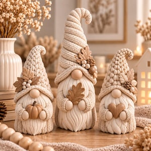 May include: Three decorative gnomes in cream and beige tones, each wearing a textured knit hat adorned with autumn leaves and berries. The left gnome holds a small pumpkin, the center gnome holds an oak leaf, and the right gnome holds a heart.