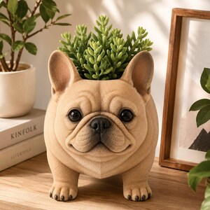 May include: A tan French bulldog-shaped planter with large dark eyes and a black nose. The dog figure has a textured surface and stands on four short legs. A green succulent plant is potted inside the top of the dog's head, filling the opening.