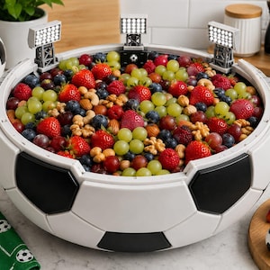 Könnte beinhalten: Eine große, runde Servierschüssel im Design eines Fußballs mit schwarzen und weißen fünfeckigen Feldern und drei am Rand befestigten Miniatur-Stadionflutlichtern, gefüllt mit einer Mischung aus frischen Erdbeeren, Weintrauben und Nüssen.