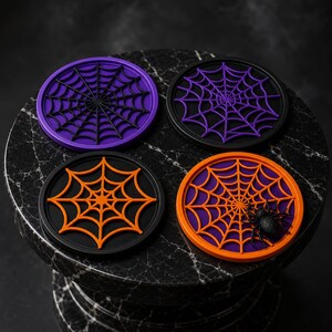 Könnte beinhalten: Vier runde Untersetzer mit erhabenen Spinnennetz-Motiven. Zwei Untersetzer haben violette Basen mit schwarzen Netzen, einer hat eine schwarze Basis mit einem orangefarbenen Netz und einer hat eine violette Basis mit orangefarbenem Netz und Spinne.